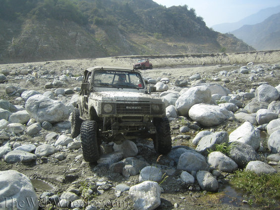 Mud-covered 1987 Suzuki Samurai driving over rocks in a dry riverbed. Another red Jeep is in the background