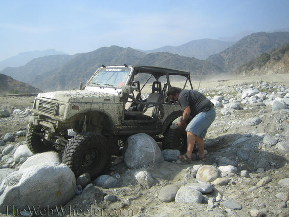 1987 Suzuki Samurai with a person adjusting the front tire in a rocky, dry riverbed with mountains in the background