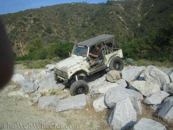 White 1987 Suzuki Samurai off-roading over large rocks, driver visible