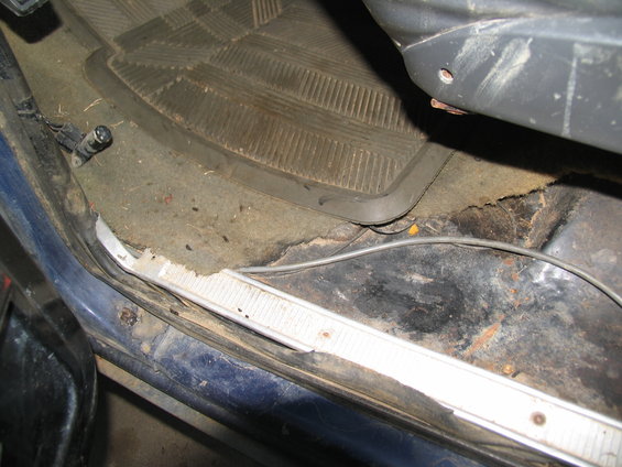 View of the driver's side floor area inside a vehicle, showing a rubber floor mat, worn carpet, and rust on the metal floor pan