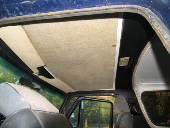 Car interior ceiling with tan carpet lining and visible damage to a sun visor. Black trim and seats are also visible
