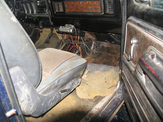 Interior of a blue vehicle, showing a worn driver's seat, rusted floor, and aftermarket radio. The dashboard has woodgrain trim