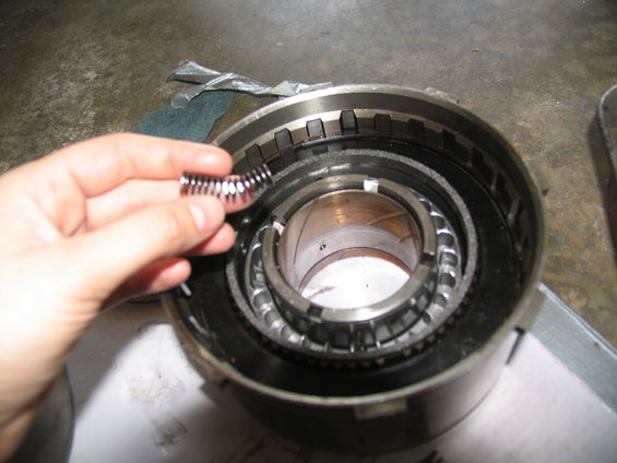 Hand holding a small spring over a disassembled transmission component with metal rings and teeth