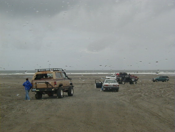 Several off-road vehicles and cars parked on a sandy beach under a cloudy sky