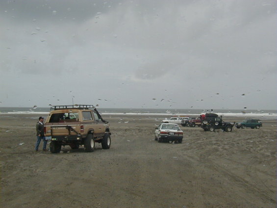 Several off-road vehicles parked on a sandy beach with the ocean in the background under a cloudy sky