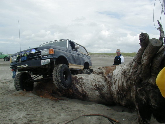 Dark blue lifted SUV with off-road tires navigating a large log on a sandy beach