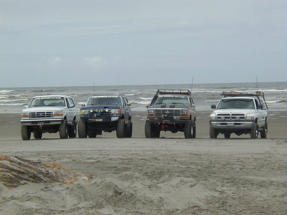 Four SUVs parked on a beach with the ocean in the background. The first SUV is white, the second is dark blue, the third is brown, and the fourth is white