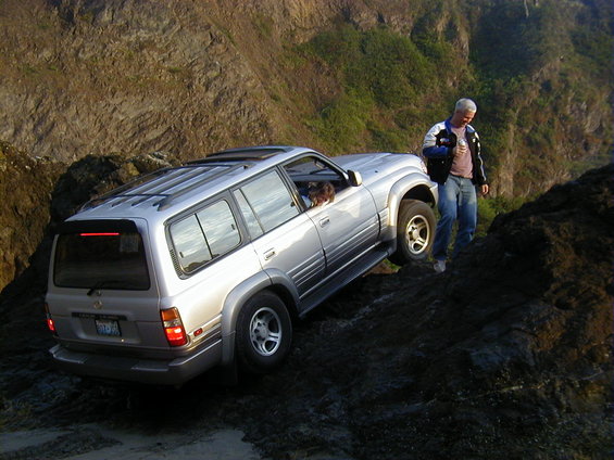 Silver SUV with roof rack driving on a rocky incline near a cliff, man standing nearby