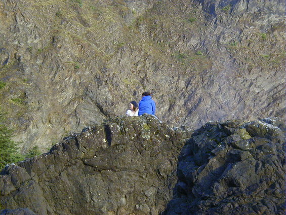 Two people sit on a rocky cliff overlooking a vast, textured rock face