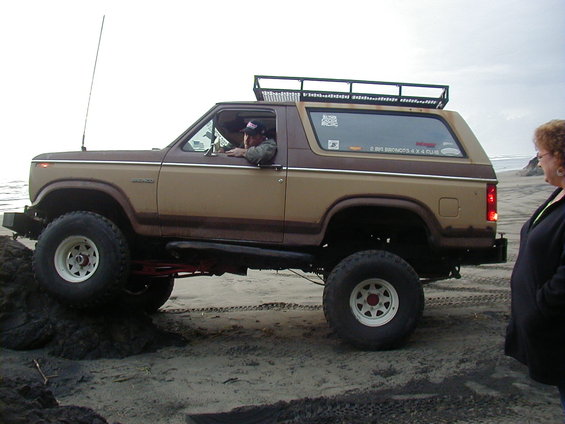 Brown and tan Ford Bronco with roof rack on sand, driver visible. A woman stands to the right