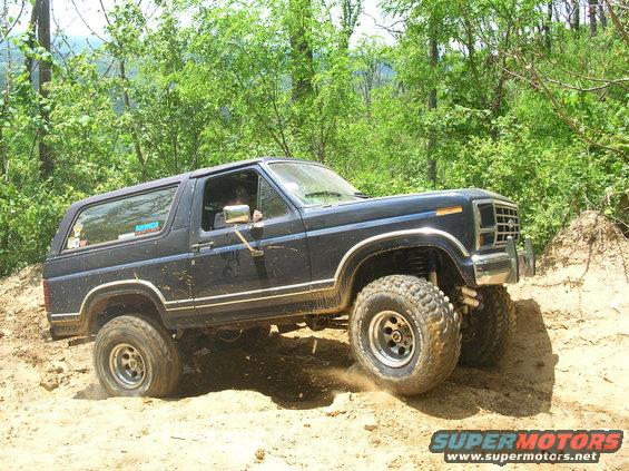 Dark blue Ford Bronco climbing a dirt hill with trees in the background. It has large off-road tires