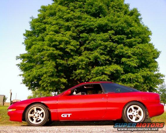 Red coupe with GT badge parked outside under a large tree