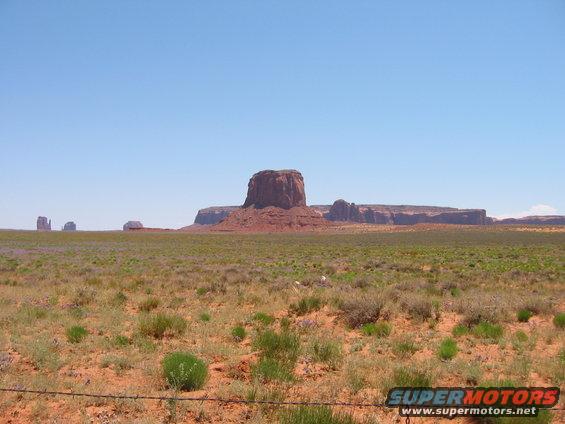 Monument Valley with mesas and desert scrub under a clear blue sky. Watermark reads SUPERMOTORS