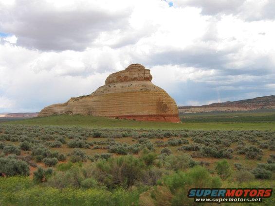 Large sandstone rock formation with layered strata under a cloudy sky, surrounded by green fields and desert brush