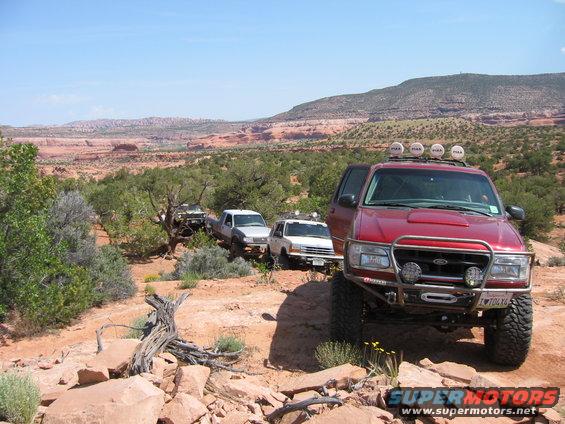 Red Ford Explorer with off-road modifications leads a convoy of trucks on a desert trail