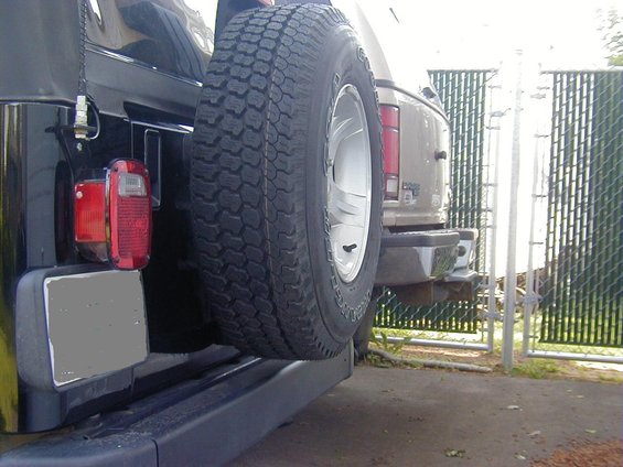 Spare tire on the back of a black Jeep Wrangler, with a tan SUV behind it