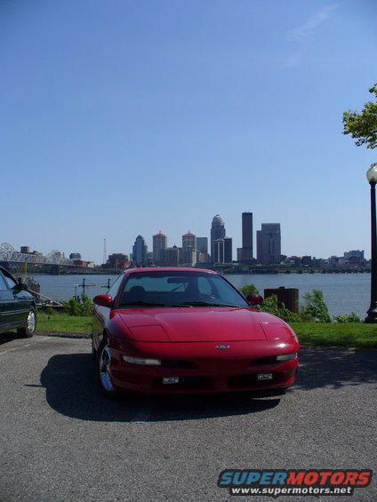 Red 1991 Ford Probe parked with a city skyline and river in the background. Another dark car is partially visible on the left