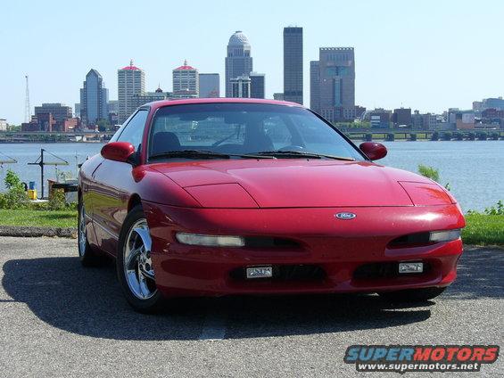 Red 1993 Ford Probe parked by a waterfront with a city skyline in the background