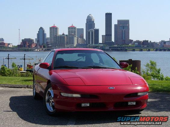 Red 1991 Ford Probe parked by a body of water with a city skyline behind it