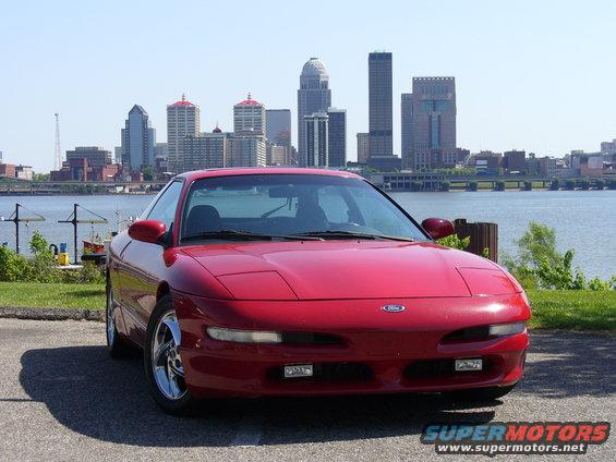 Red Ford Probe with chrome wheels parked by a waterfront, city skyline in background