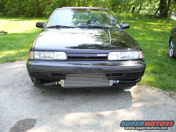 Front view of a black car with a large intercooler installed below the bumper. Mazda logo visible on grille. Another car partially visible on the right