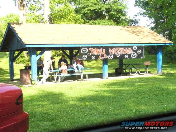 People sit under a blue and brown picnic shelter with a banner reading 'MDY MAZDAS' and multiple Mazda logos