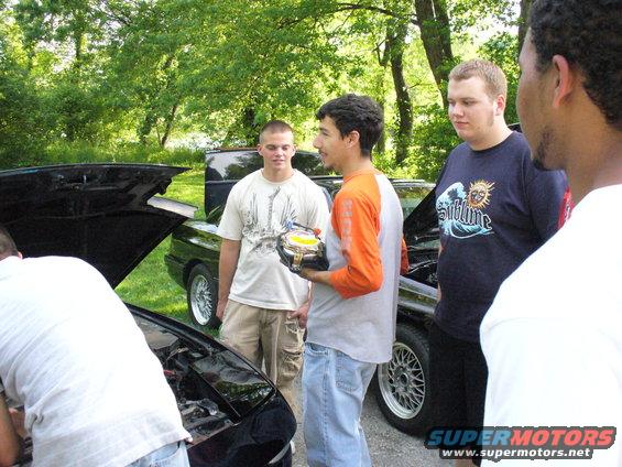 Three men examine car engines with hoods open; one holds a car part