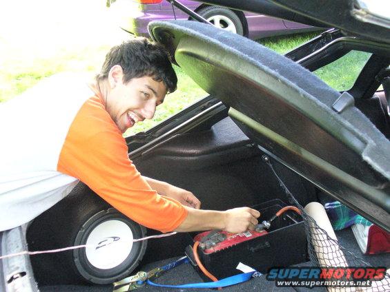 Person connecting battery terminals in car trunk, with subwoofer visible. Another car is partially visible in the background