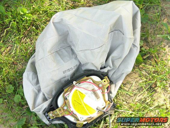 Deactivated airbag module with yellow warning label and part number UT11573-01D, lying on grass