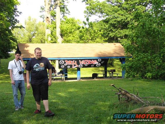 Two guys stand in a grassy area near a covered picnic shelter. Graffiti on the shelter reads 'MDY MAZDAS' with Mazda logos