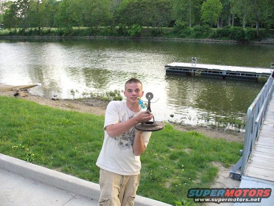 Man holding a metal sculpture made of car parts by a lake