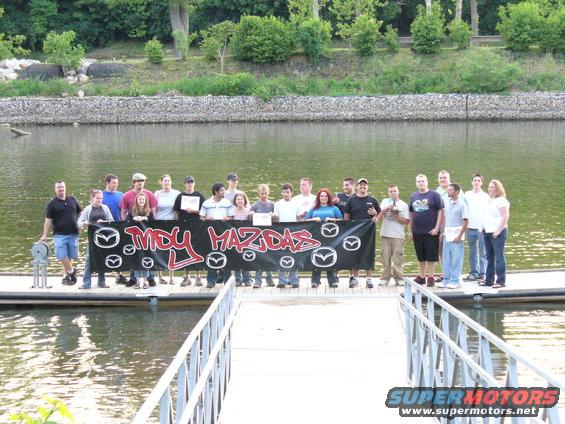 Group of people holding "MDY MAZDAS" banner by a lake