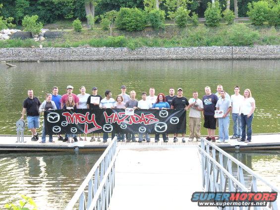 Group of people on a dock holding a banner that says "MDY MAZDAS" with Mazda logos