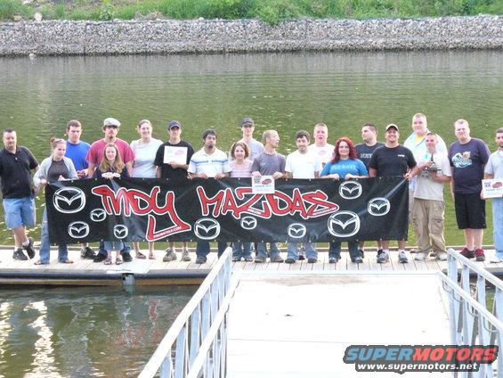 Group of people on a dock holding a black banner with red text and Mazda logos