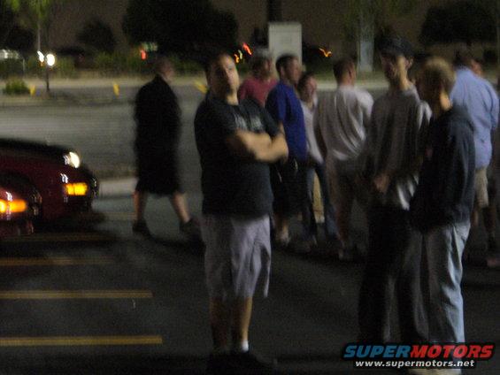 Red car front corner with headlights on, parked next to a group of people