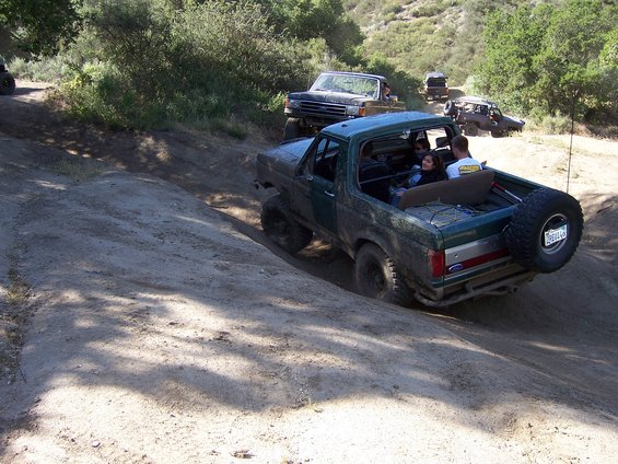 Green Ford Bronco driving up a dirt hill with people inside. Another Bronco and a pickup truck are behind it