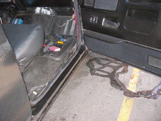 Car interior with black seats, tools on floor mat, and a puddle of dark liquid outside the open door