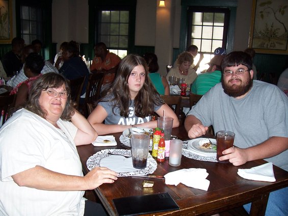 Woman, teen girl, and man sit at a table in a restaurant. Condiments and drinks are on the table