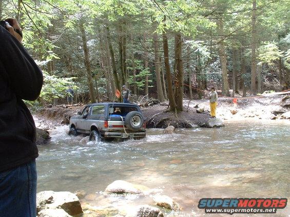 Ford Bronco driving through a stream with trees and people on the bank