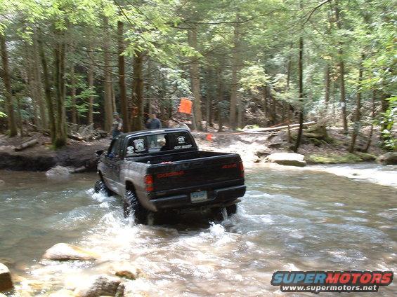 Black Dodge Ram pickup truck with "4x4" decal on tailgate driving through stream. "DODGE" lettering visible on tailgate