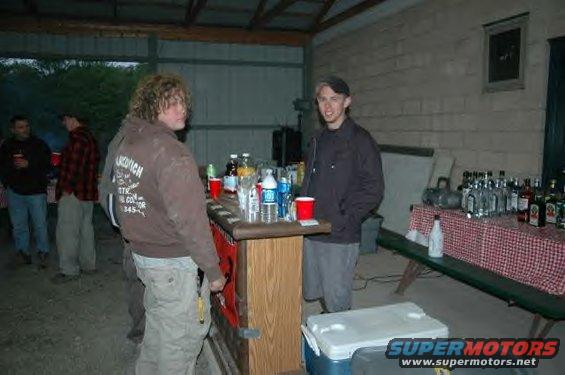 People stand around a bar with drinks and bottles on a table behind it