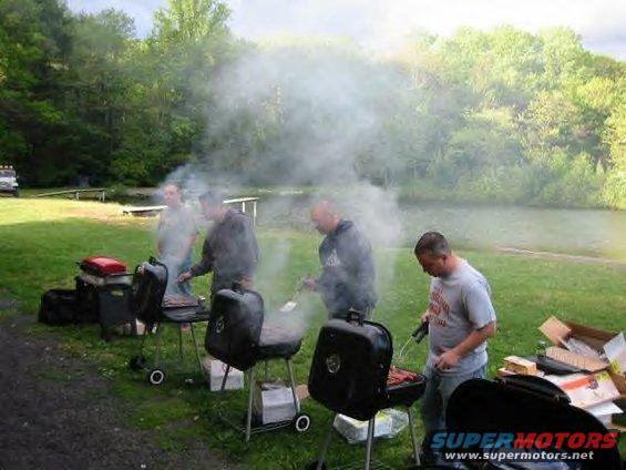 People grilling outdoors with smoke rising, near a body of water and trees