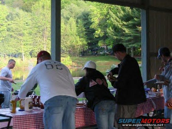 People gathered around a picnic table laden with food and drinks, with trees and a body of water in the background