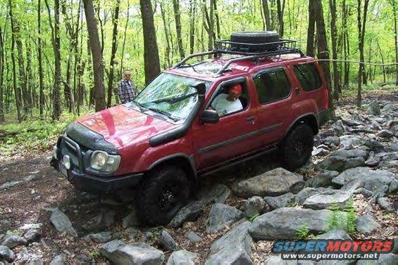 Red SUV with roof rack and spare tire navigating a rocky forest trail. A person stands behind it