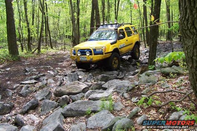 Yellow SUV with blue bumper and roof rack driving on a rocky forest trail