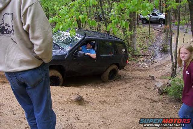 Black SUV stuck in mud with a white truck further up a trail and people standing around