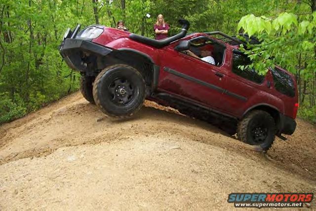 Red SUV with snorkel and off-road tires climbing a dirt hill, two people visible in background