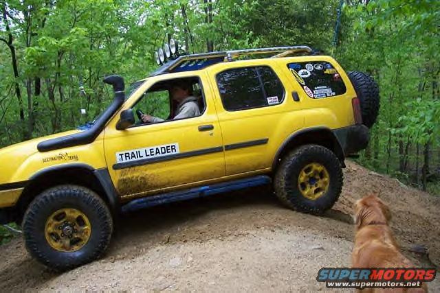 Yellow SUV with snorkel, roof rack, and spare tire on the back, driving on a dirt trail