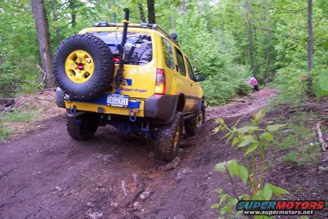 Yellow SUV with spare tire on rear, driving on a dirt trail. Another vehicle is visible in the distance