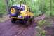 1991 Ford Bronco - A yellow SUV navigates a muddy trail in a wooded area, with another vehicle seen further up the path.